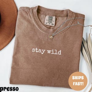 May include: Espresso-colored t-shirt with the words "stay wild" in white lettering. A silver necklace with a small pendant rests on the shirt. A brown hat is visible in the upper left corner. The shirt is folded and has a tag.