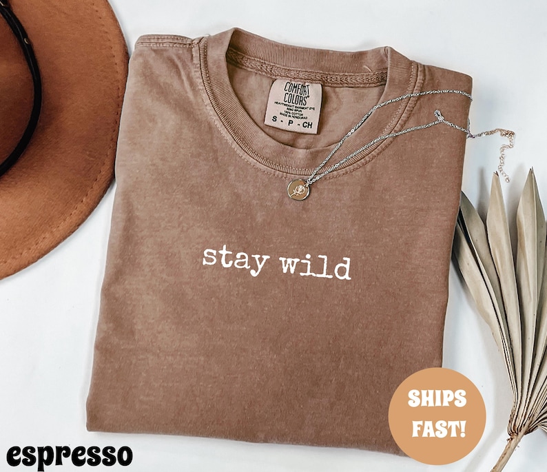 May include: Espresso-colored t-shirt with the words "stay wild" in white lettering. A silver necklace with a small pendant rests on the shirt. A brown hat is visible in the upper left corner. The shirt is folded and has a tag.
