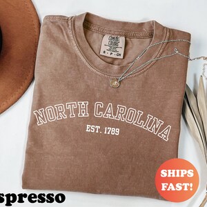 Puede incluir: Camiseta color espresso con el texto blanco "NORTH CAROLINA EST. 1789". Un collar plateado con un colgante descansa sobre la camiseta. La camiseta tiene cuello redondo y una peque&ntilde;a etiqueta. Un c&iacute;rculo naranja con "SHIPS FAST!" es visible.