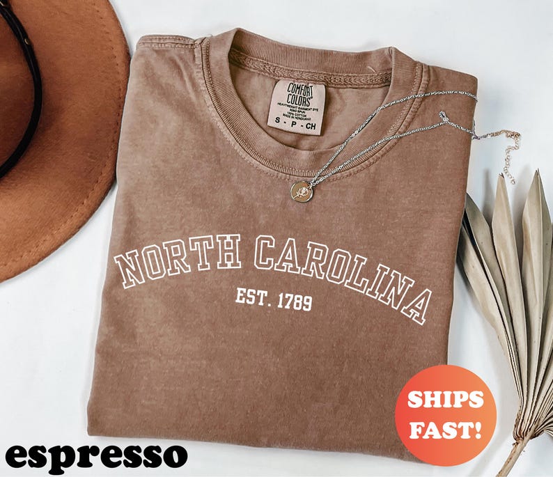 Puede incluir: Camiseta color espresso con el texto blanco "NORTH CAROLINA EST. 1789". Un collar plateado con un colgante descansa sobre la camiseta. La camiseta tiene cuello redondo y una peque&ntilde;a etiqueta. Un c&iacute;rculo naranja con "SHIPS FAST!" es visible.