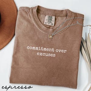May include: Espresso-colored t-shirt with the phrase "commitment over excuses" in white lettering. The shirt has a crew neck and short sleeves. A silver necklace with a small pendant is draped across the shirt. The shirt is made by Comfort Colors.
