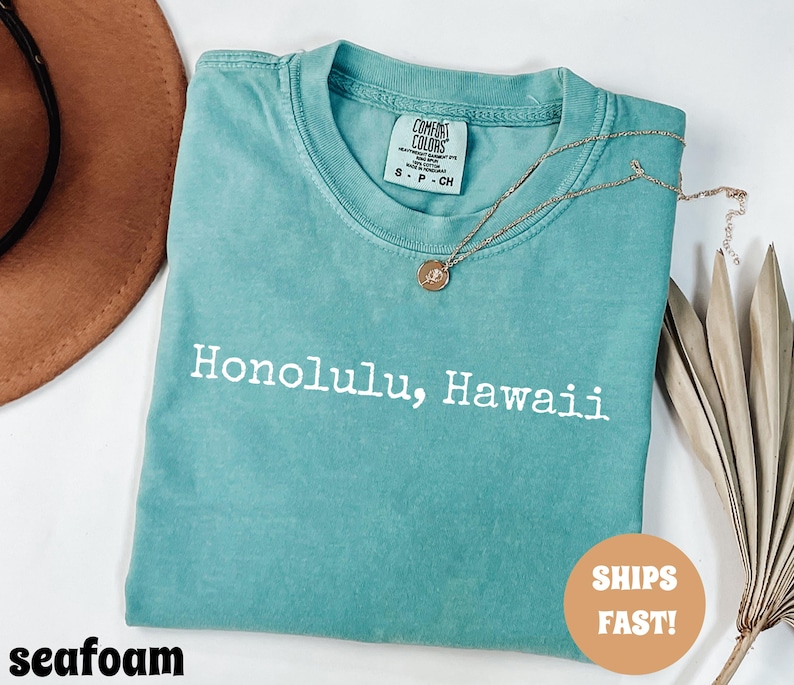 Może przedstawiać: Miętowa koszulka z białym napisem "Honolulu, Hawaii". Koszulka ma okrągły dekolt i kr&oacute;tkie rękawy. Złoty naszyjnik z wisiorkiem jest zarzucony na koszulkę. Koszulka jest złożona i ma metkę Comfort Colors.