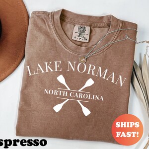 May include: Espresso-colored t-shirt with the text "Lake Norman North Carolina" and crossed oars in white. A silver necklace with a pendant rests on the shirt. The shirt has a Comfort Colors tag.