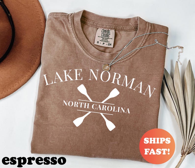 May include: Espresso-colored t-shirt with the text "Lake Norman North Carolina" and crossed oars in white. A silver necklace with a pendant rests on the shirt. The shirt has a Comfort Colors tag.