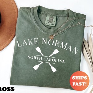 May include: A moss green t-shirt with the text "LAKE NORMAN NORTH CAROLINA" and crossed oars in white. The shirt has a crew neck and a small tag. A gold necklace and a round orange sticker that says "SHIPS FAST!" are also visible.