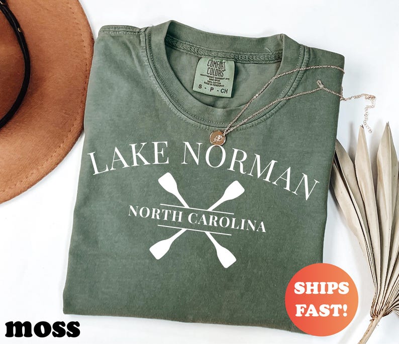 May include: A moss green t-shirt with the text "LAKE NORMAN NORTH CAROLINA" and crossed oars in white. The shirt has a crew neck and a small tag. A gold necklace and a round orange sticker that says "SHIPS FAST!" are also visible.