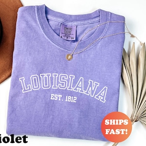May include: A violet-colored t-shirt with the word "LOUISIANA" in white block letters and "EST. 1812" underneath. The shirt is made by Comfort Colors. A gold necklace with a pendant is draped across the shirt. A round orange sticker says "SHIPS FAST!"