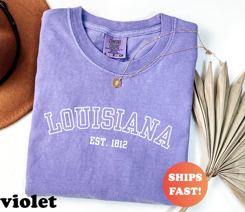 May include: A violet-colored t-shirt with the word "LOUISIANA" in white block letters and "EST. 1812" underneath. The shirt is made by Comfort Colors. A gold necklace with a pendant is draped across the shirt. A round orange sticker says "SHIPS FAST!"