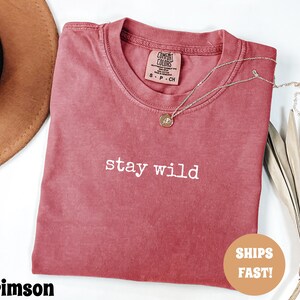 May include: Crimson-colored t-shirt with the words "stay wild" printed in white. The shirt has a crew neck and a small tag on the collar. A gold necklace with a pendant rests on the shirt. A brown hat is visible in the background.