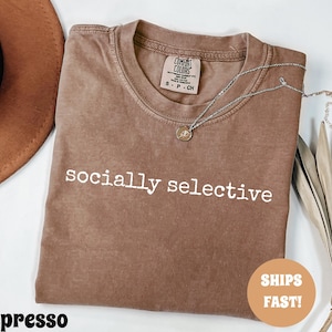 May include: Espresso-colored t-shirt with the words "socially selective" printed in white. A silver necklace with a small pendant rests on the shirt. The shirt is folded, and a brown hat is visible in the background.