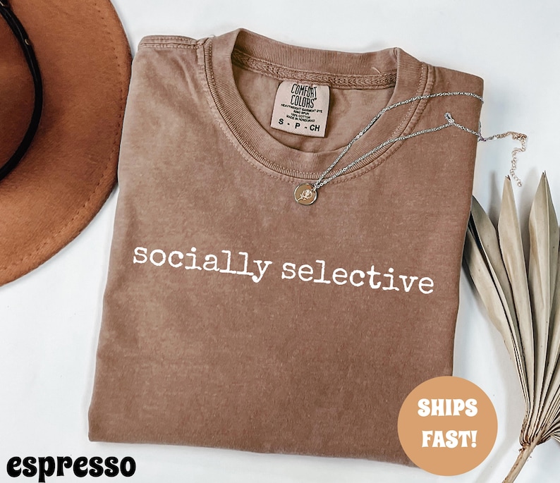 May include: Espresso-colored t-shirt with the words "socially selective" printed in white. A silver necklace with a small pendant rests on the shirt. The shirt is folded, and a brown hat is visible in the background.