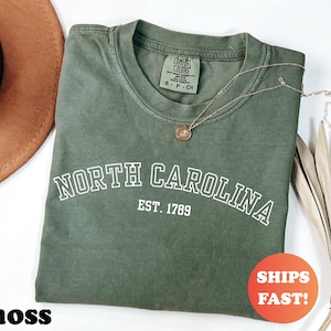 Puede incluir: Una camiseta verde musgo con las palabras "NORTH CAROLINA" en letras blancas y "EST. 1789" debajo. Un collar dorado con un colgante descansa sobre la camiseta. La camiseta tiene una etiqueta Comfort Colors. El texto "SHIPS FAST!" est&aacute; en un c&iacute;rculo rojo.