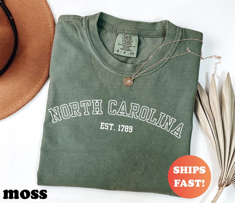 Puede incluir: Una camiseta verde musgo con las palabras "NORTH CAROLINA" en letras blancas y "EST. 1789" debajo. Un collar dorado con un colgante descansa sobre la camiseta. La camiseta tiene una etiqueta Comfort Colors. El texto "SHIPS FAST!" est&aacute; en un c&iacute;rculo rojo.