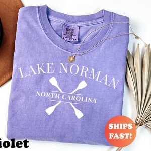 May include: A violet Comfort Colors t-shirt with the text "Lake Norman North Carolina" and crossed oars in white. A gold necklace and a dried palm leaf are on the right. A brown hat is on the left. An orange sticker says "Ships Fast!"