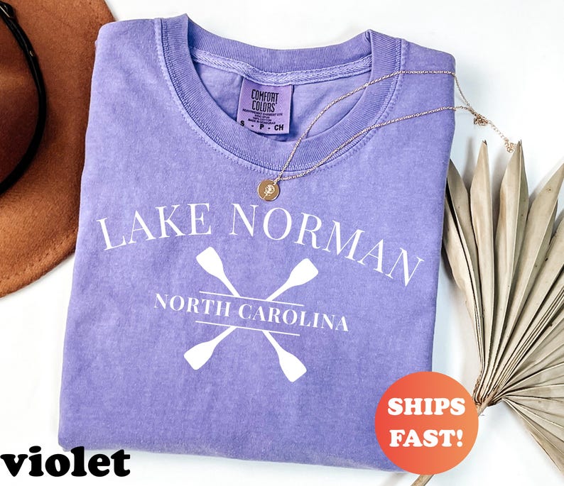 May include: A violet Comfort Colors t-shirt with the text "Lake Norman North Carolina" and crossed oars in white. A gold necklace and a dried palm leaf are on the right. A brown hat is on the left. An orange sticker says "Ships Fast!"