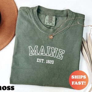 Könnte beinhalten: Ein moosgrünes T-Shirt mit dem Wort "MAINE" in weißen Blockbuchstaben und "EST. 1820" darunter. Eine goldene Halskette mit Anhänger liegt auf dem Shirt. Das Shirt hat einen Rundhalsausschnitt und ein kleines Etikett. Ein orangefarbener Kreis mit "SHIPS FAST!" befindet sich im Bild.
