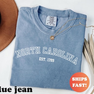 Puede incluir: Una camiseta azul vaquero con el texto "NORTH CAROLINA EST. 1789" en blanco. Un collar de oro con un colgante est&aacute; sobre la camiseta. La camiseta tiene una etiqueta Comfort Colors. Una pegatina que dice "SHIPS FAST!" est&aacute; en la camiseta.