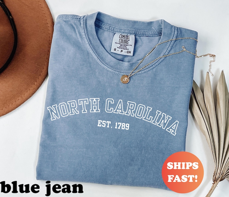 Puede incluir: Una camiseta azul vaquero con el texto "NORTH CAROLINA EST. 1789" en blanco. Un collar de oro con un colgante est&aacute; sobre la camiseta. La camiseta tiene una etiqueta Comfort Colors. Una pegatina que dice "SHIPS FAST!" est&aacute; en la camiseta.