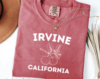 May include: Crimson-colored t-shirt with the words "IRVINE CALIFORNIA" in white block letters. A graphic of oranges and leaves is below the word "IRVINE". A gold necklace rests on the shirt. The shirt has a Comfort Colors tag.