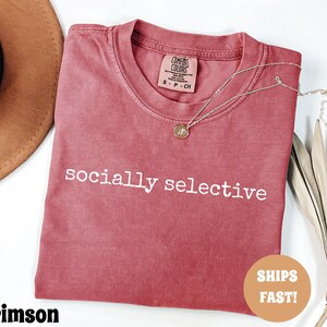 May include: Crimson-colored t-shirt with the words "socially selective" printed in white. The shirt has a crew neck and a small tag on the collar. A gold necklace with a pendant is draped across the shirt. A brown hat is visible in the background.