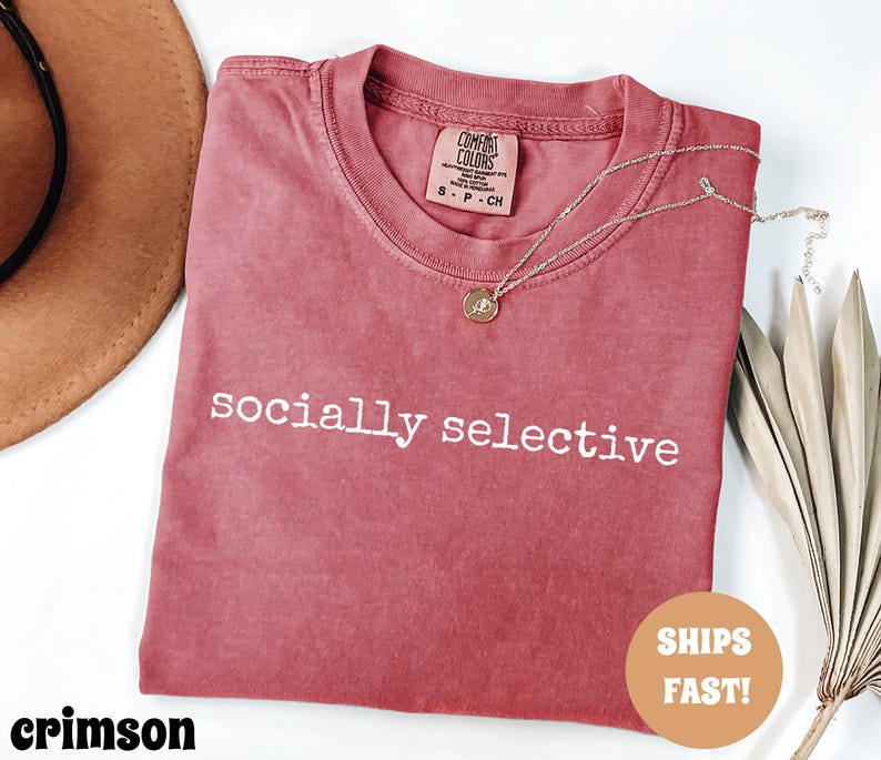 May include: Crimson-colored t-shirt with the words "socially selective" printed in white. The shirt has a crew neck and a small tag on the collar. A gold necklace with a pendant is draped across the shirt. A brown hat is visible in the background.