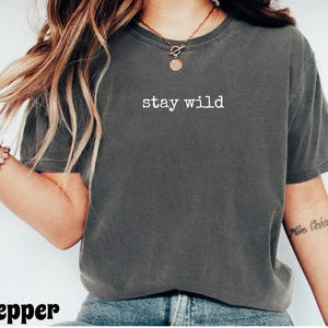 May include: A dark gray t-shirt with the words "stay wild" printed in white. The shirt has short sleeves and a crew neck. The shirt is made of a soft, comfortable material.