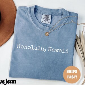 Może przedstawiać: Niebieska koszulka dżinsowa z białym napisem "Honolulu, Hawaii". Koszulka ma okrągły dekolt i małą metkę na g&oacute;rze plec&oacute;w. Złoty naszyjnik z wisiorkiem jest zarzucony na koszulkę. Koszulka jest złożona.