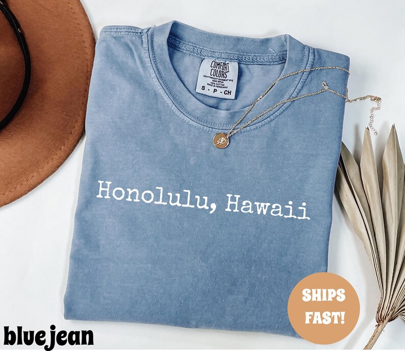 Może przedstawiać: Niebieska koszulka dżinsowa z białym napisem "Honolulu, Hawaii". Koszulka ma okrągły dekolt i małą metkę na g&oacute;rze plec&oacute;w. Złoty naszyjnik z wisiorkiem jest zarzucony na koszulkę. Koszulka jest złożona.