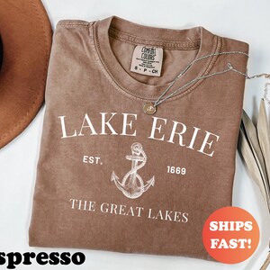 May include: Espresso-colored t-shirt with the text "LAKE ERIE" and an anchor graphic. The shirt also includes the text "EST. 1669" and "THE GREAT LAKES". A silver necklace with a pendant is draped across the shirt.