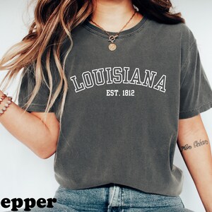 May include: A dark gray t-shirt with the word "LOUISIANA" in white, arched text, and "EST. 1812" below it. The shirt is a casual style, likely made of cotton, and suitable for everyday wear. The word "pepper" is at the bottom of the image.