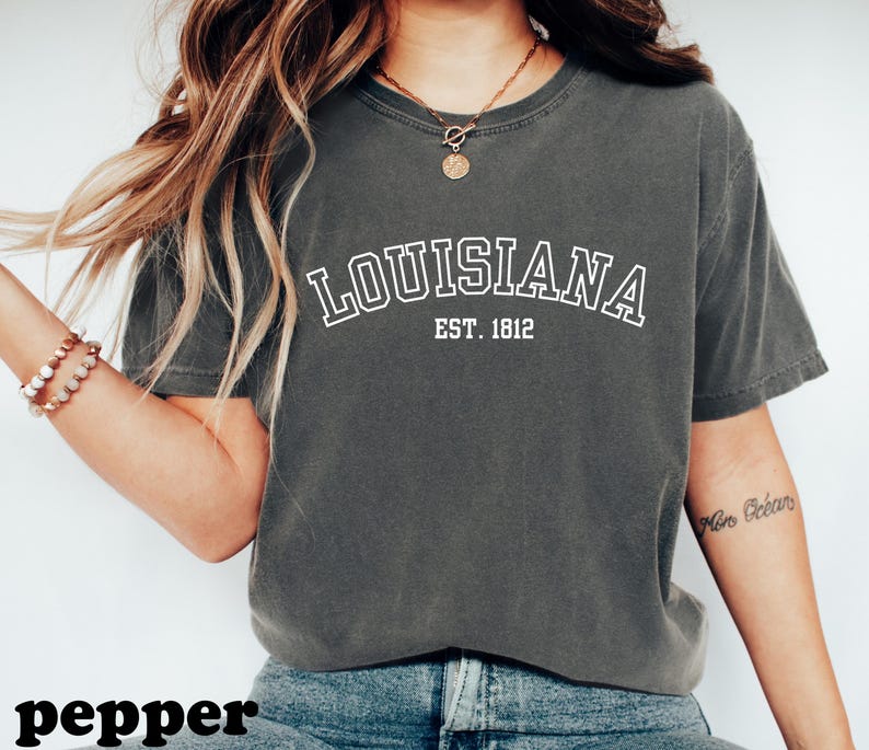May include: A dark gray t-shirt with the word "LOUISIANA" in white, arched text, and "EST. 1812" below it. The shirt is a casual style, likely made of cotton, and suitable for everyday wear. The word "pepper" is at the bottom of the image.