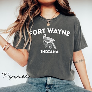 Może przedstawiać: Ciemnoszara koszulka z białym napisem "FORT WAYNE INDIANA". Pomiędzy tekstem znajduje się szczegółowa ilustracja kardynała. Koszulka ma swobodny styl.