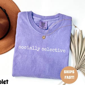 May include: A violet-colored Comfort Colors t-shirt with the phrase "socially selective" printed in white. A gold necklace with a pendant is draped on the shirt. A brown hat is in the background.