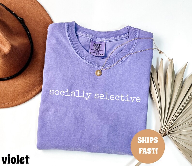 May include: A violet-colored Comfort Colors t-shirt with the phrase "socially selective" printed in white. A gold necklace with a pendant is draped on the shirt. A brown hat is in the background.