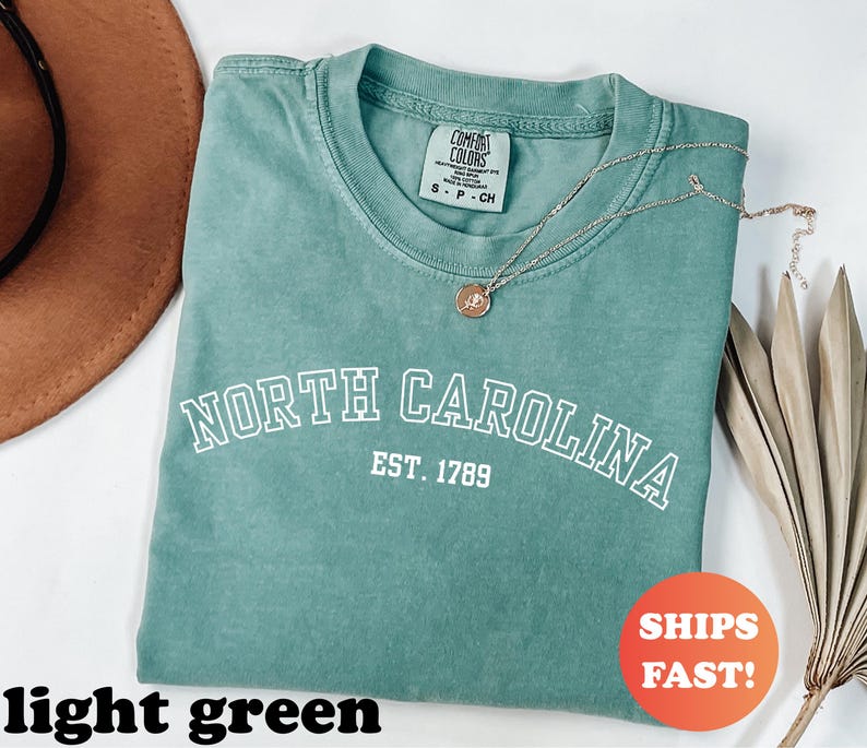 Puede incluir: Una camiseta verde claro con las palabras "NORTH CAROLINA" y "EST. 1789" impresas en blanco. Un collar dorado con un colgante descansa sobre la camiseta. La camiseta est&aacute; doblada y se muestra con un sombrero marr&oacute;n y una planta seca.