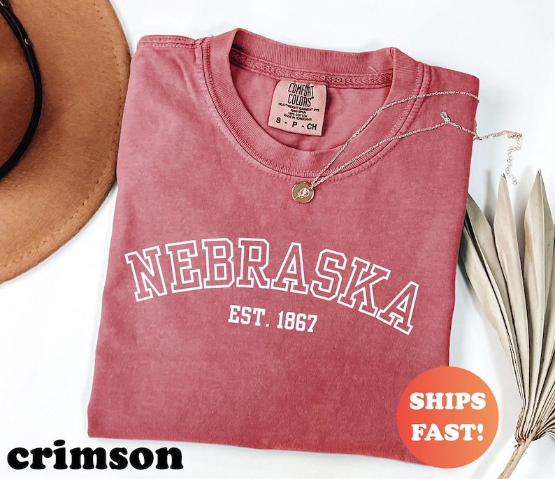 May include: Crimson-colored t-shirt with the word "NEBRASKA" in white block letters and "EST. 1867" underneath. The shirt has a crew neck and a small tag. A gold necklace with a pendant is draped across the shirt.
