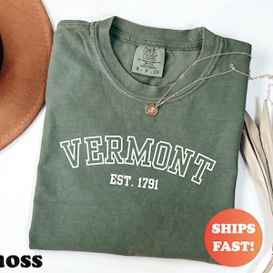May include: A moss green t-shirt with the word "VERMONT" in white block letters and "EST. 1791" underneath. The shirt has a crew neck and a small tag on the collar. A gold necklace with a pendant is draped across the shirt.