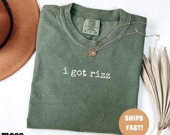 May include: A moss green t-shirt with the text "i got rizz" in white. A gold necklace with a pendant rests on the shirt. The shirt has a crew neck and short sleeves. A "Ships Fast!" sticker is visible.