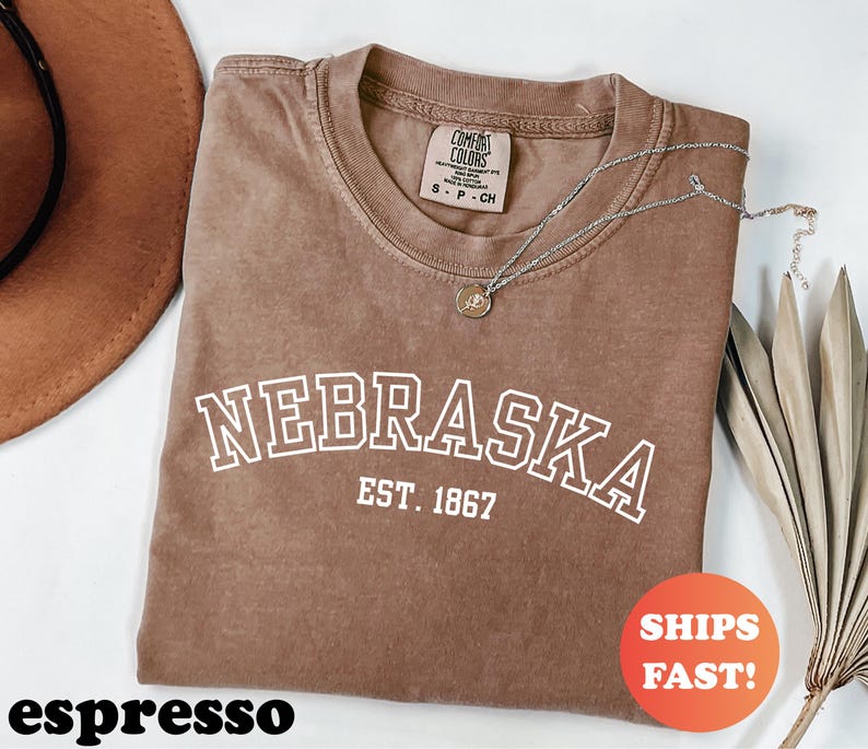 May include: Espresso-colored t-shirt with the word "NEBRASKA" in white block letters, arched above "EST. 1867." A silver necklace with a pendant rests on the shirt. The shirt is folded, with a brown hat and dried leaves in the background.