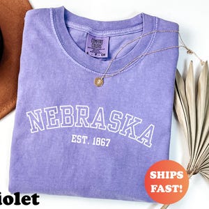 May include: A violet-colored t-shirt with the word "NEBRASKA" in white block letters and "EST. 1867" underneath. A gold necklace with a pendant rests on the shirt. A brown hat and dried leaves are in the background. A sticker says "SHIPS FAST!"