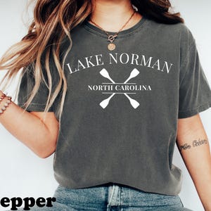 May include: A dark gray t-shirt with the text "Lake Norman North Carolina" in white, along with crossed oars. The word "pepper" is at the bottom of the image. The shirt is a casual style.