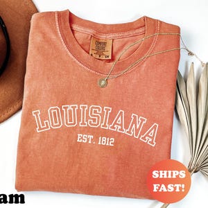 May include: A coral-colored t-shirt with the word "LOUISIANA" in white block letters, with "EST. 1812" below. A gold necklace and a brown hat are visible. A round orange sticker says "SHIPS FAST!"