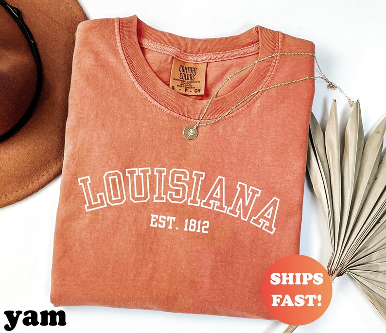 May include: A coral-colored t-shirt with the word "LOUISIANA" in white block letters, with "EST. 1812" below. A gold necklace and a brown hat are visible. A round orange sticker says "SHIPS FAST!"