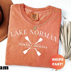 May include: A coral-colored t-shirt with the text "Lake Norman North Carolina" and crossed oars in white. The shirt has a crew neck and a small tag. A gold necklace and a "Ships Fast!" sticker are also visible.