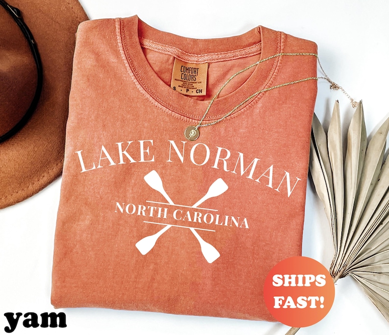 May include: A coral-colored t-shirt with the text "Lake Norman North Carolina" and crossed oars in white. The shirt has a crew neck and a small tag. A gold necklace and a "Ships Fast!" sticker are also visible.