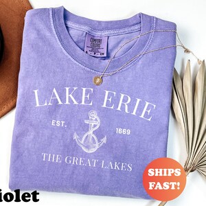 May include: A violet-colored t-shirt with the text "LAKE ERIE" and an anchor graphic. The shirt also includes the text "EST. 1669" and "THE GREAT LAKES". A gold necklace and a brown hat are also in the image.