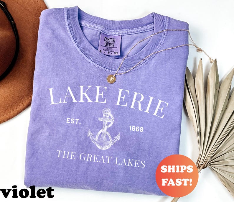 May include: A violet-colored t-shirt with the text "LAKE ERIE" and an anchor graphic. The shirt also includes the text "EST. 1669" and "THE GREAT LAKES". A gold necklace and a brown hat are also in the image.