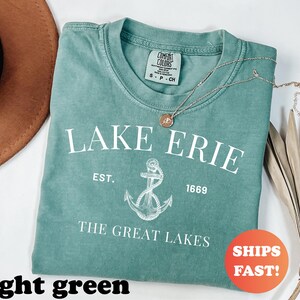 May include: A light green t-shirt with the text "LAKE ERIE" and an anchor graphic. The shirt also includes the text "EST. 1669" and "THE GREAT LAKES". A gold necklace with a pendant rests on the shirt.