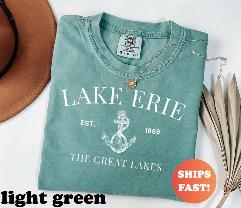 May include: A light green t-shirt with the text "LAKE ERIE" and an anchor graphic. The shirt also includes the text "EST. 1669" and "THE GREAT LAKES". A gold necklace with a pendant rests on the shirt.