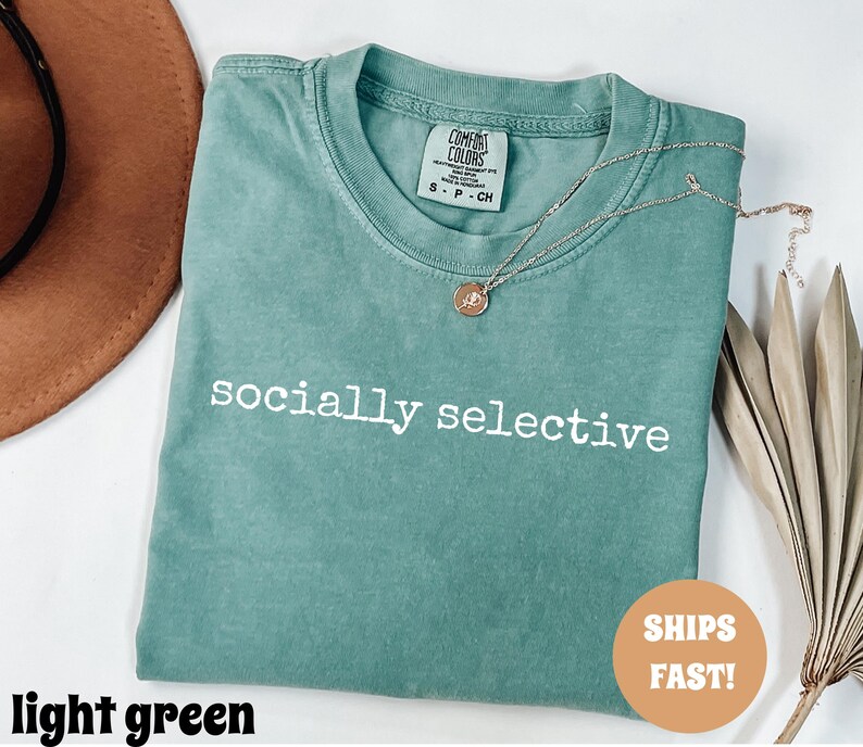 May include: A light green t-shirt with the words "socially selective" printed in white. The shirt has a crew neck and short sleeves. A gold necklace with a pendant is draped across the shirt.